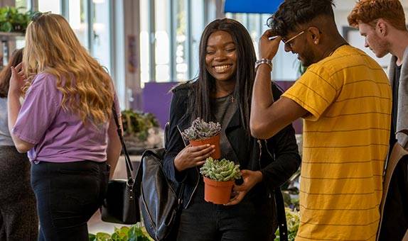 Three students at Penrhyn Road campus Students' Union. A girl is facing the camera smiling holding two cactuses in plant pots 